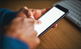 A man's hands tap the blank screen of a cellphone. A computer keyboard can be seen in the background.